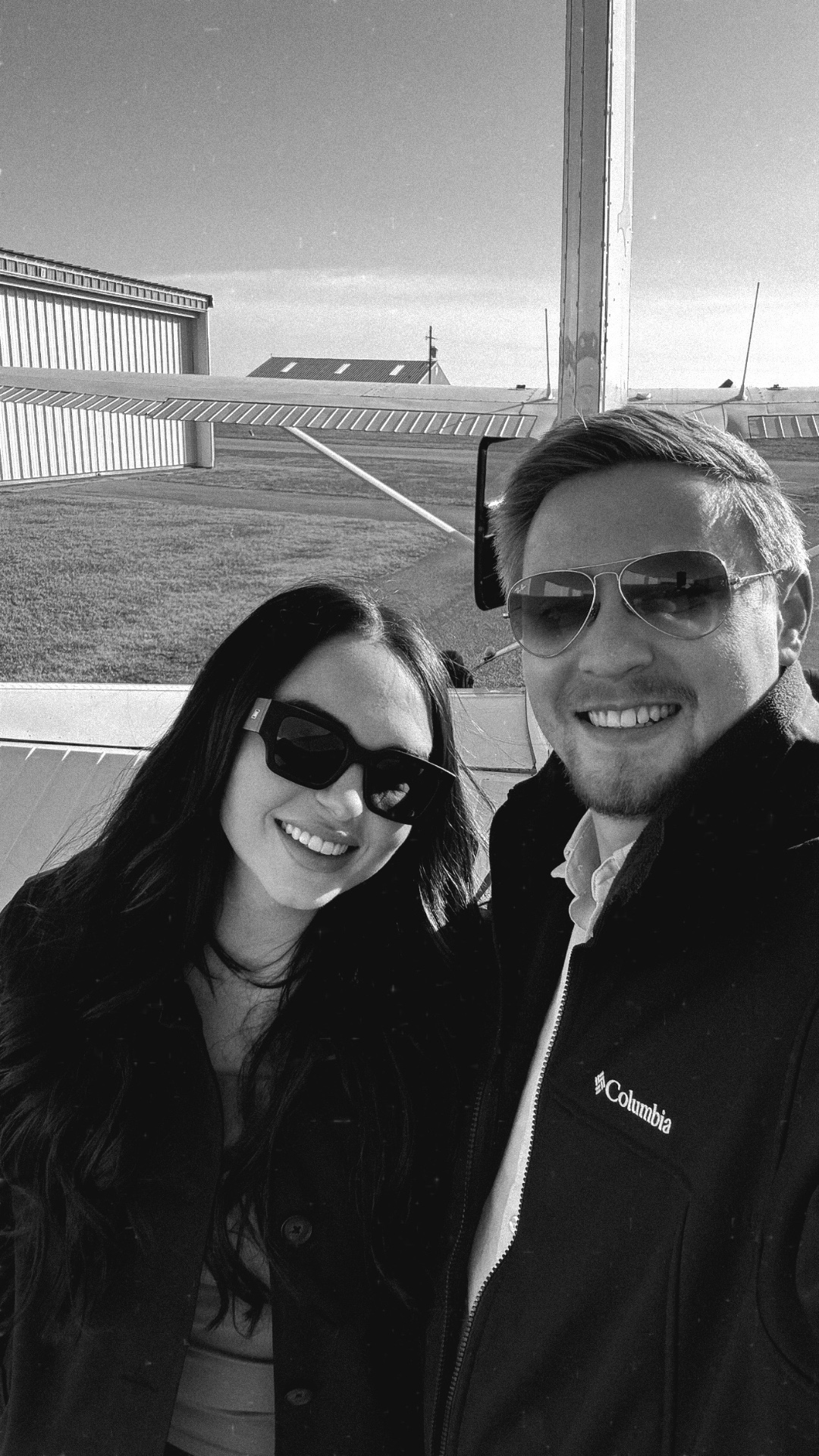 Black and white selfie of a couple at the airfield by a small aircraft